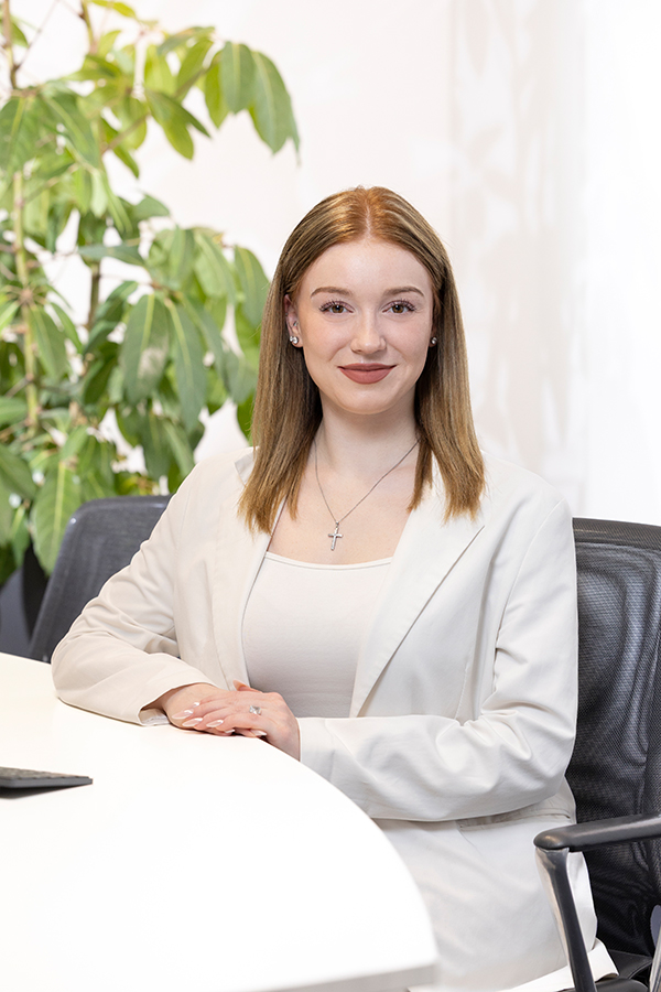 Ausbildung bei der MIZ. Junge Frau in weißem Blazer sitzt lächelnd an einem Tisch.